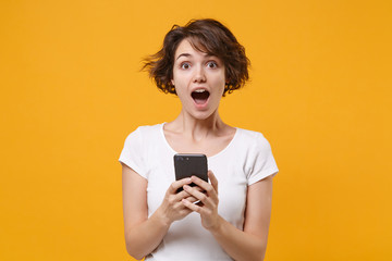 Shocked young brunette woman girl in white t-shirt posing isolated on yellow orange background studio portrait. People lifestyle concept. Mock up copy space. Using mobile phone, typing sms message.