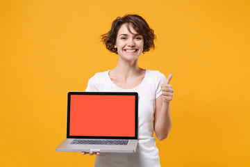 Smiling young brunette woman girl in white t-shirt posing isolated on yellow background. People lifestyle concept. Mock up copy space. Hold laptop pc computer with blank empty screen showing thumb up.