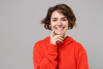 Cheerful young brunette woman girl in casual red hoodie posing isolated on grey background studio portrait. People sincere emotions lifestyle concept. Mock up copy space. Put hand prop up on chin.
