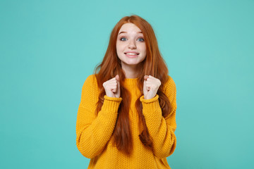 Excited young redhead woman girl in yellow sweater posing isolated on blue turquoise background studio portrait. People lifestyle concept. Mock up copy space. Clenching fists, wait for special moment.