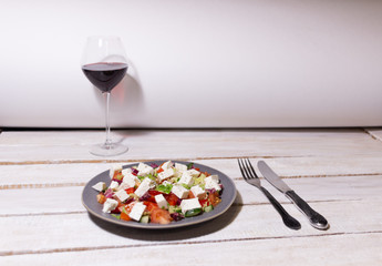 plate with greek salad, fork and knife, red wine glass on white wooden table