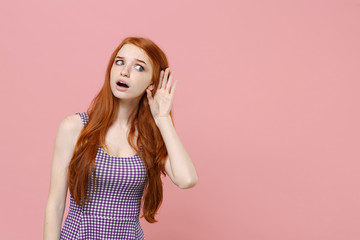 Shocked young redhead woman girl in plaid dress posing isolated on pastel pink background studio portrait. People emotions lifestyle concept. Mock up copy space. Try to hear you with hand gesture.