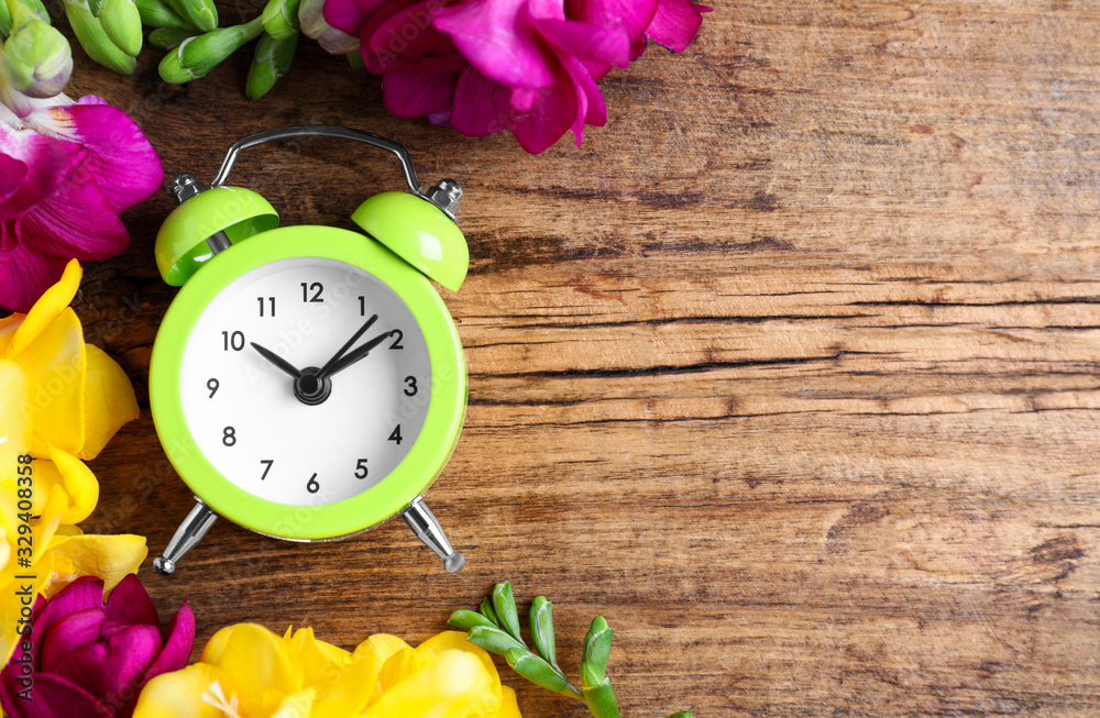 Poster green alarm clock with spring flowers and space for text on wooden table, flat lay. time change
