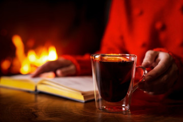 Woman with tasty mulled wine reading book near fireplace at home, closeup