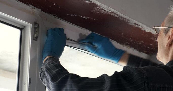 Professional repairman in blue gloves and a gray shirt carefully removes the plastic seal from the white window with a tool. repair in a new apartment. close-up camera movement