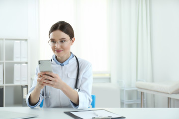 Young female doctor with smartphone at table in office