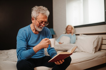 Portrait of smiling good looking elderly man with grey hair and beard drinking coffee and reading book while sitting on bed in bedroom.