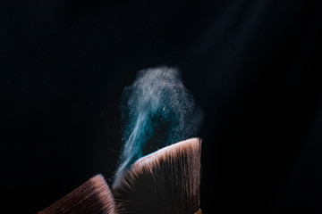 Two make up brushes with flying color powder, product on black background. Concept of glamour make up in minimal black