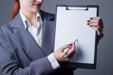 Closeup studio portrait, business woman in a gray business suit, jacket. holding in his hand a folder with a blank sheet, a pencil pointing to free space for text. On a gray background.