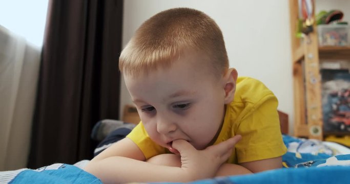 Boy of preschool age lies in bed on his stomach, bites nails on hand, looks at tablet screen, does not take his eyes off, props cheek with hand, close-up, children's room, home furnishings, childhood