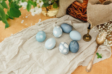Modern Easter eggs on rustic table with spring flowers, basket, linen cloth. Stylish pastel blue Easter eggs painted in natural dye from red cabbage. Happy Easter. Rural still life