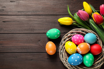 Colorful Easter eggs in decorative nest and tulips on wooden background, flat lay. Space for text