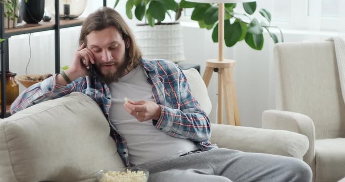 Man talking on mobile phone and eating popcorn while relaxing on sofa in the living room