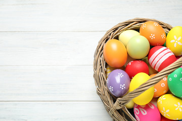 Colorful Easter eggs in basket on white wooden table, top view. Space for text