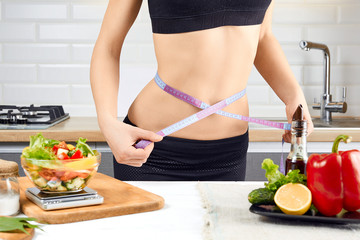 Obraz na płótnie Canvas Young sports girl with naked torso measuring her waist with measuring tape in the kitchen. Diet and weigh loss concept