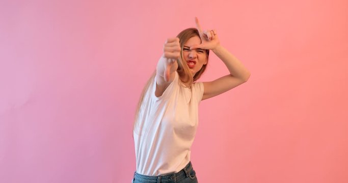 the girl shows the gesture of a loser or a loser , in a white t- shirt with a grin on her face, on a pink background