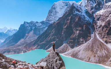 A man standing on the rock in front of turquoise Chola lake and  Tabuche foothills, from the way to Dzongla, Sagarmatha national park, Everest Base Camp 3 Passes Trek, Nepal