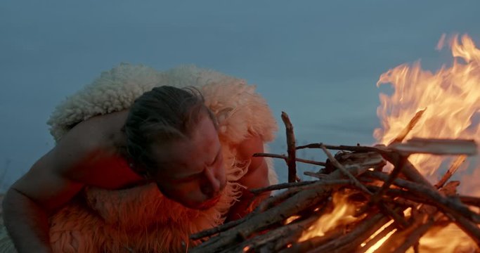 Man in animal hide blowing at burning fire then warming hands in evening in barbarian camp - slow motion