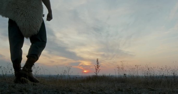 Man with sword and animal hide walking in meadow then stopping and looking at sunset sky in nature
