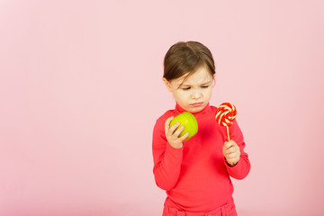 Little girl chooses between a lollipop and a green apple. The concept of proper nutrition. A child in a pink studio holds a sweet of sugar in his hand and an apple. Difficulty of choice