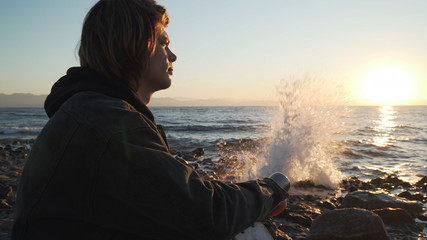 Young caucasian boy drink hot tea from thermos at seashore at sunset