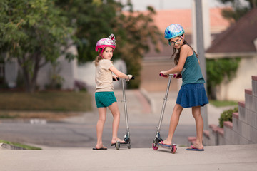 Girls are wearing safety helmets while scootering on the sidewalk of their LA city.