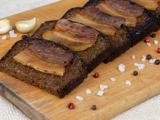 Black bread toasts with bacon on wooden background with garlic and salt