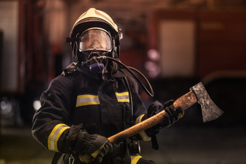 Fototapeta premium female firefighter portrait wearing full equipment, oxygen mask, and an axe. smoke and fire trucks in the background.