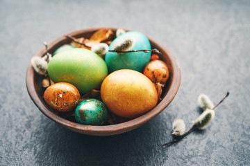 Bright easter eggs in a clay bowl and fluffy willow branches on a dark background, soft focus. 