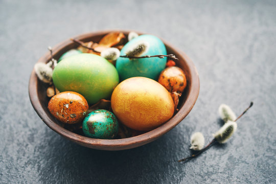 Bright easter eggs in a clay bowl and fluffy willow branches on a dark background, soft focus. 