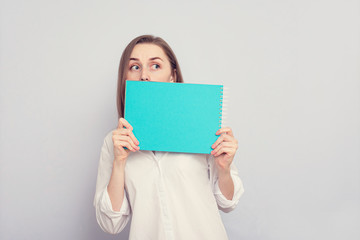 Fototapeta premium Scared student girl hides behind a notebook, grey background, copy space