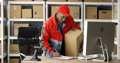 African American mailman in uniform working at computer in post office store with parcels. Postman scanning bar code on carton box with scanner, registering it and filling in invoice document.