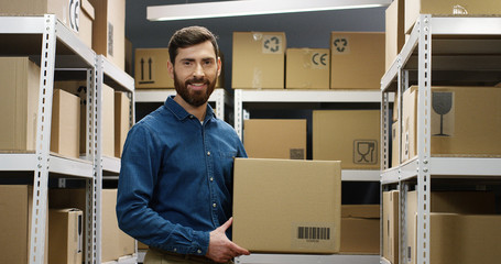 Portrait of handsome young male courier in smiling cheerfully to camera in postal storage of parcels. Postman taking parcel carton box from shelf at working place.