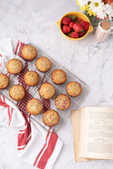 Top View of Homemade Strawberry Muffins on Cooling Rack on White Marble Countertop; Old Cookbook Nearby, Red and White Kitchen Towel; Strawberries and Flowers on Counter