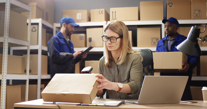 Beautiful Caucasian woman post worker sitting at desk and working at computer in postal delivery store. Female scanning parcel and registering it in system in post office. Male couriers on background.