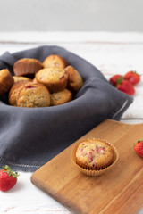 Bowl of Freshly Baked Homemade Strawberry Muffins in a Wood Bowl; One Isolated on Wood Cutting Board in Front; Fresh Strawberrries Scattered Around; White Wood Tabletop