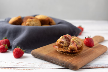 Bowl of Freshly Baked Homemade Strawberry Muffins in a Wood Bowl; One Isolated on Wood Cutting Board in Front; Fresh Strawberrries Scattered Around; White Wood Tabletop