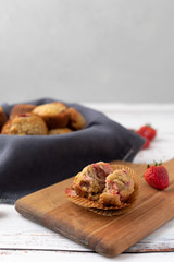 Bowl of Freshly Baked Homemade Strawberry Muffins in a Wood Bowl; One Isolated on Wood Cutting Board in Front; Fresh Strawberrries Scattered Around; White Wood Tabletop