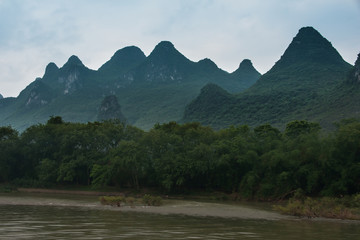 Guilin, China - May 10, 2010: Along Li River. Landscape with green forested karst mountain under blueish cloudscape. Brown water and dark tree-filled shoreline up front.
