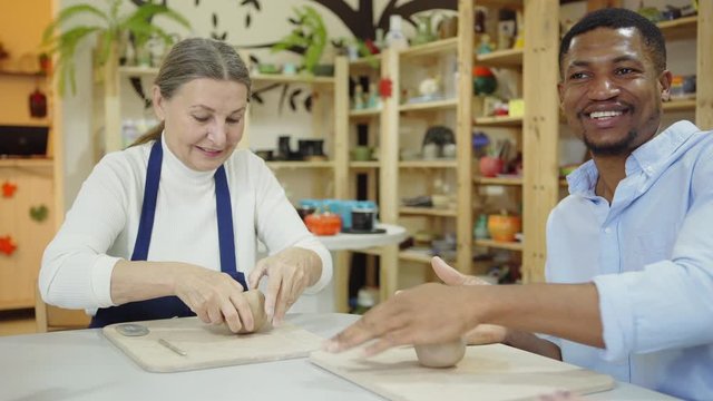 Tracking medium shot of middle aged ceramic artist teaching elderly Caucasian woman and young African man how to wedge clay sitting at desk in art studio. People enjoying talking at work