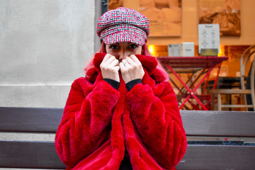 Mujer sentada  en un banco  con una chaqueta de pelos roja y gorra  a conjunto