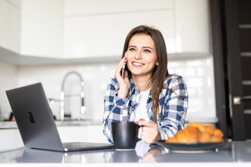 Young woman using her laptop and talking on phone during breakfast in the kitchen at home