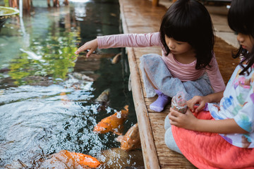 two little girls having fun feeding koi fish by the pond