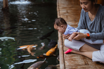 asian mother and her baby daughter see koi fish from the edge of a pond