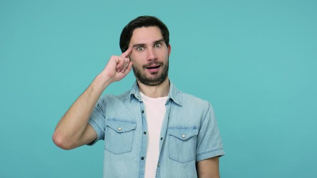 I'm crazy! Bearded guy in jeans shirt with awkward dumb grimace showing stupid gesture, having insane idea, being out of mind, making cuckoo silly face. indoor studio shot isolated on blue background