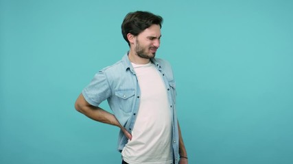 Backache. Tired unhealthy male worker in jeans shirt touching injured sore back, suffering lower lumbar discomfort, muscle pain of overwork, pinched nerve. studio shot isolated on blue background