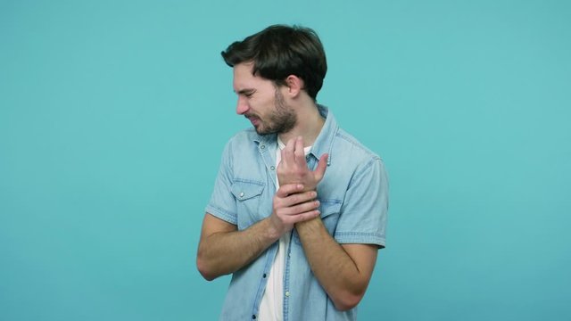Hand damage, injury. Bearded guy in jeans shirt massaging sore wrist, suffering arm sprain, carpal tunnel syndrome or pinched nerve, medical problem. indoor studio shot isolated on blue background