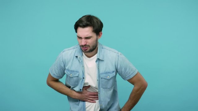 Digestion problems. Bearded guy in jeans shirt feeling sudden stomach pain, severe abdominal distress, suffering constipation, gastritis, pancreatic diseases. studio shot isolated on blue background