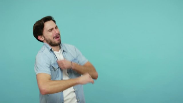 Lively active bearded guy in jeans shirt dancing admiring favorite song with quick rhythm, listening to energetic club music, celebrating success. indoor studio shot isolated on blue background