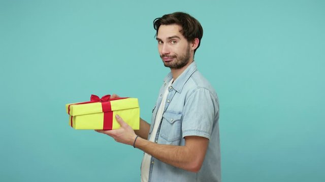 Nice bearded man unwrapping gift and expressing disappointment, showing thumbs down, dislike to bad present, guy dissatisfied with birthday surprise. indoor studio shot isolated on blue background